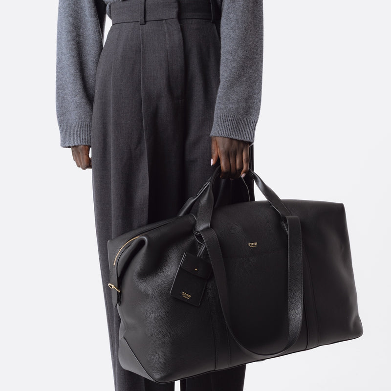 Model holding STOW London's Weekend bag in Black pebbled leather featuring detachable pouch and gold-coated stainless steel hardware on a white background
