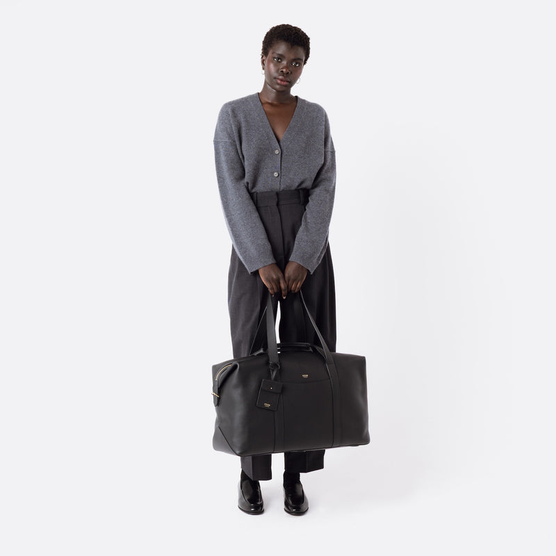 Model holding STOW London's Weekend bag in Black pebbled leather featuring detachable pouch and gold-coated stainless steel hardware on a white background
