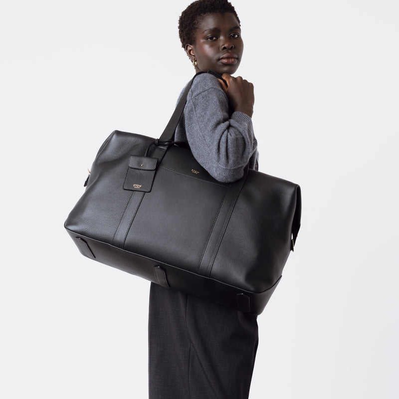 Model wearing STOW London's Weekend bag in Black pebbled leather on her shoulder featuring detachable pouch and gold-coated stainless steel hardware on a light grey background