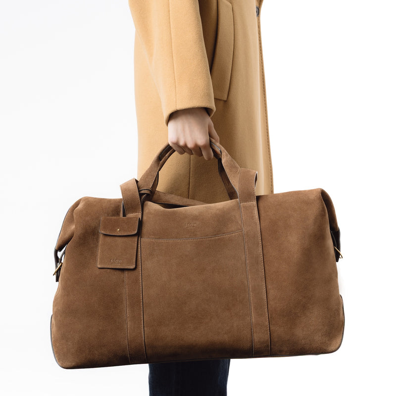 Model holding STOW London's Weekend bag in Chocolate suede leather at her side on a white background