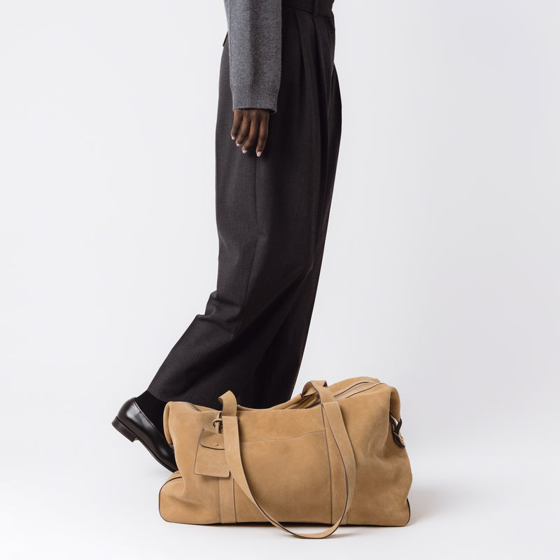 STOW London's Weekend bag in Soft Sand suede set down at model's feet on a light grey background 