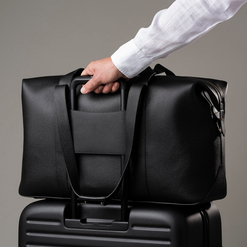 STOW Leather Weekend Bag in Black pebbled leather being pulled on top of wheeled luggage by model.