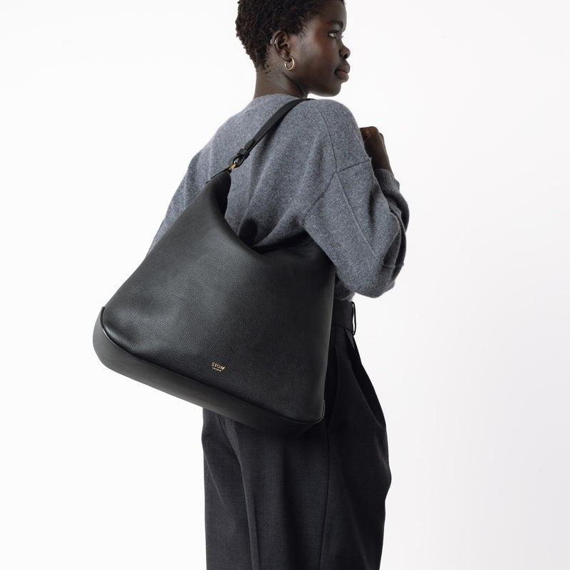 Model wearing STOW London's Maxi Curve bag in Black pebbled leather on her shoulder featuring gold-coated stainless steel hardware on a light grey background