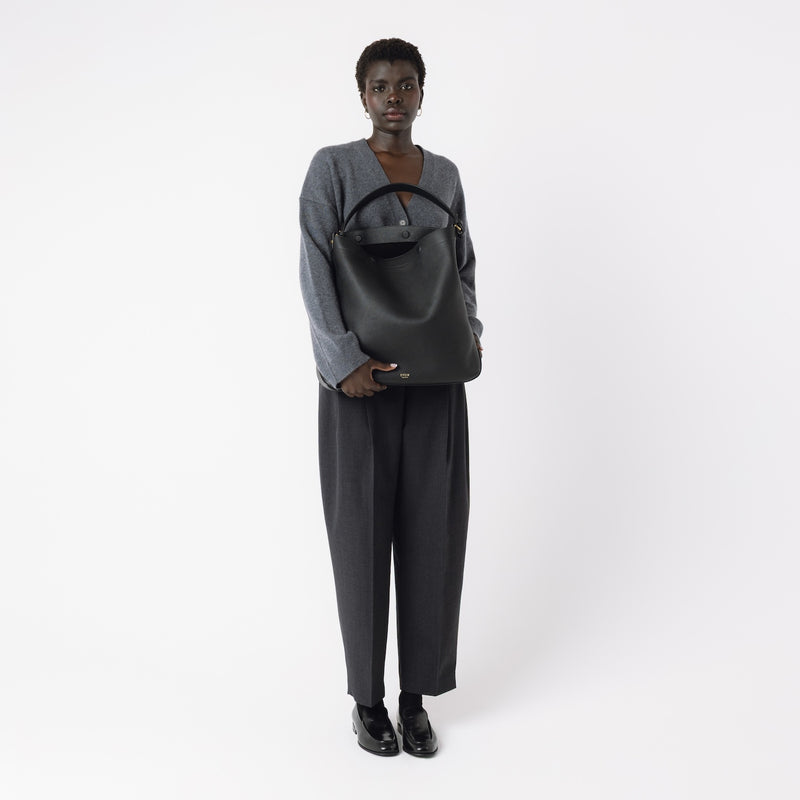 Model holding STOW London's Maxi Curve bag in Black pebbled leather featuring gold-coated stainless steel hardware on a light grey background