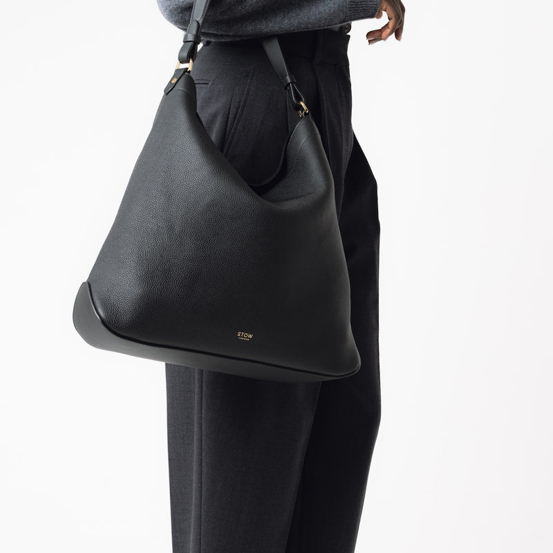 Model wearing STOW London's Maxi Curve bag in Black pebbled leather on her arm featuring gold-coated stainless steel hardware on a light grey background