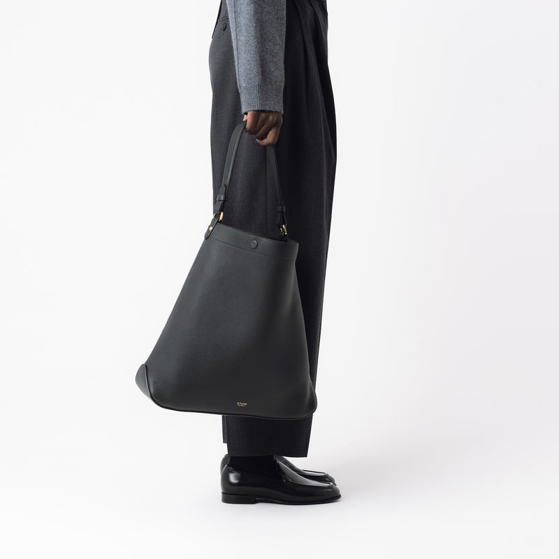 Model holding STOW London's Maxi Curve bag in Black pebbled leather at her side in an open bucket bag style featuring gold-coated stainless steel hardware on a light grey background