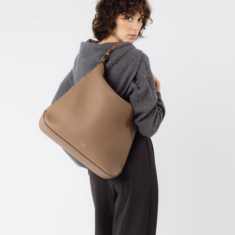 Model wearing STOW London's Maxi Curve bag in Mocha pebbled leather on her shoulder featuring gold-coated stainless steel hardware on a light grey background