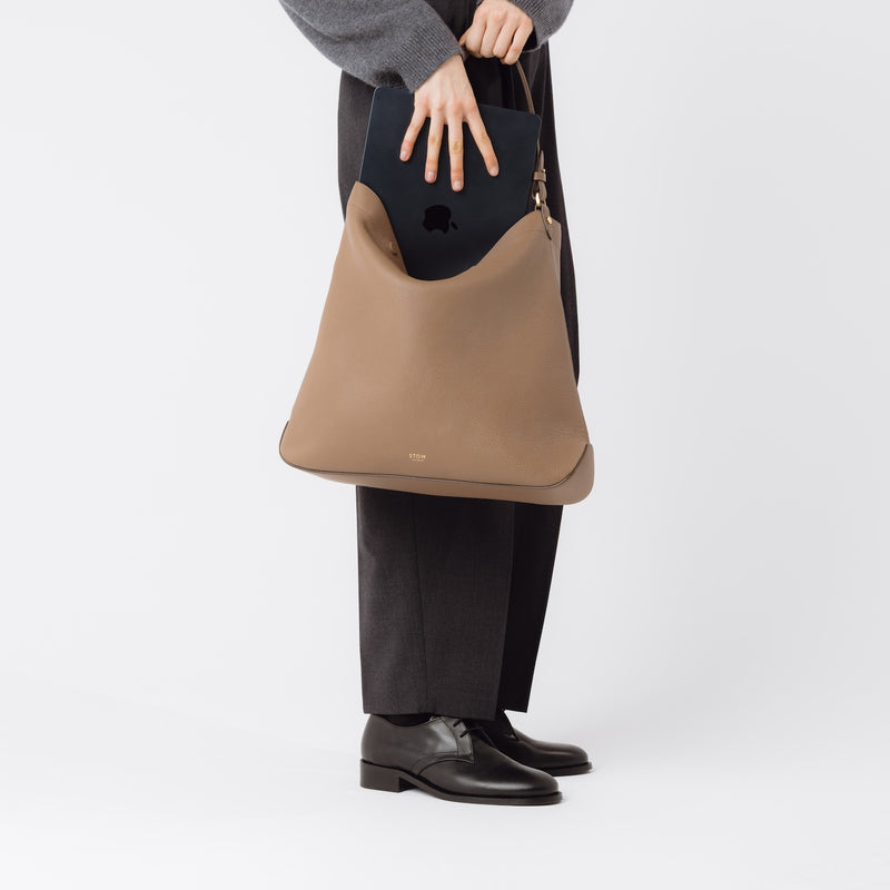 Model putting a laptop into STOW London's Maxi Curve bag in Mocha pebbled leather on a light grey background