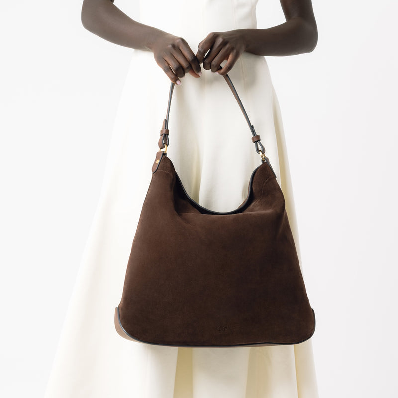 Model holding STOW London's Maxi Curve bag in Chocolate suede leather featuring gold-coated stainless steel hardware on a light grey background