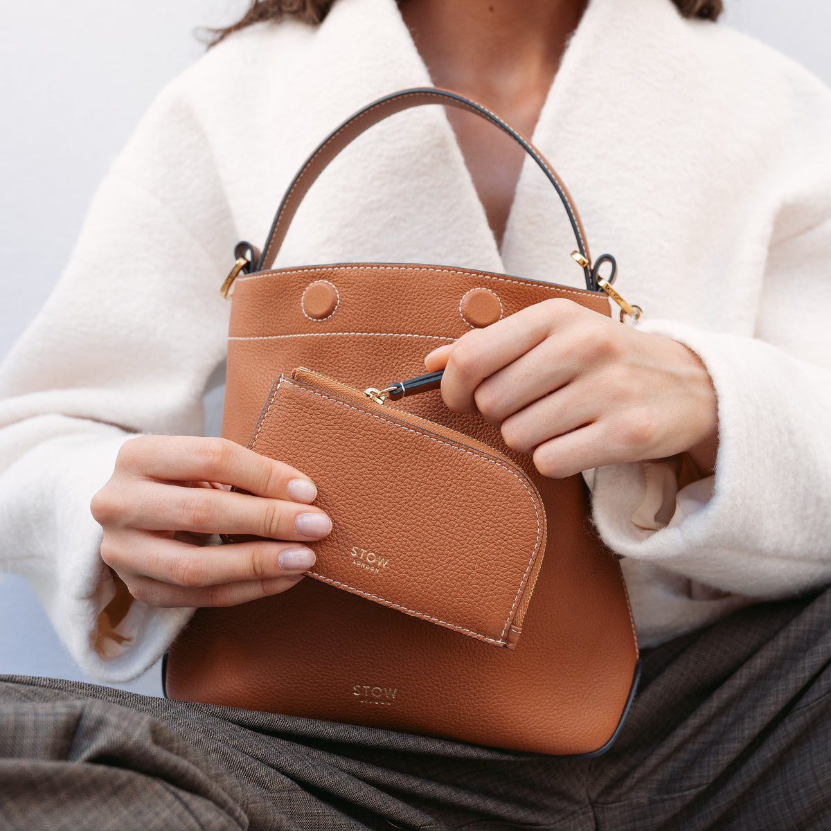 Model holding STOW London's Zip Wallet and Midi Curve bag in Earth Tan pebbled leather featuring a small gold embossed logo on the front 
